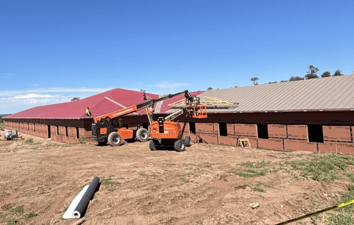 Construction site showing two orange boom lifts working on roofing a large building with red and gray metal roofs under a clear blue sky.