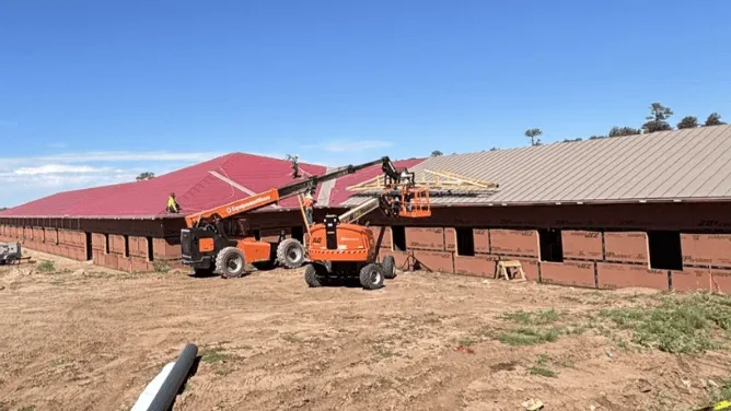 Construction site showing two orange boom lifts working on roofing a large building with red and gray metal roofs under a clear blue sky.